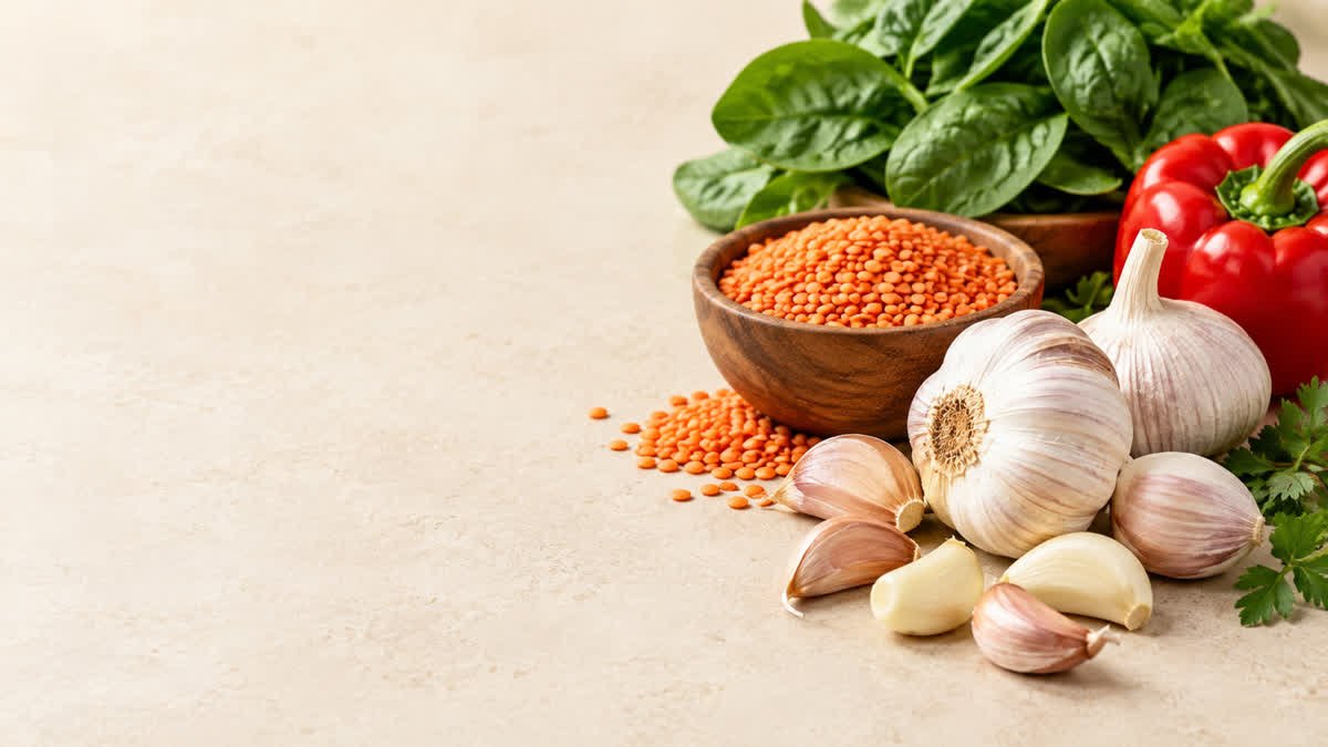 Fresh garlic bulbs and cloves beside lentils and leafy greens in a nutrition-focused food arrangement.