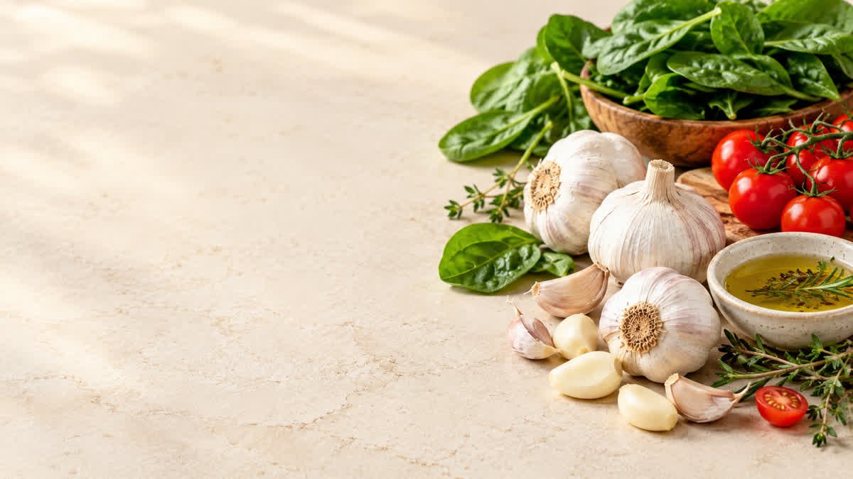 Fresh garlic bulbs and cloves beside leafy greens and tomatoes in a healthy food arrangement.