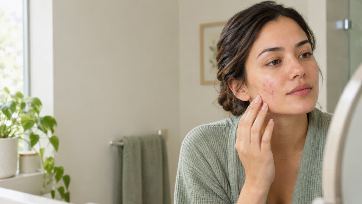 Adult woman examining mild acne blemishes on her cheek in a bathroom mirror, with natural skin texture, soft daylight, and a neutral beige and sage background.