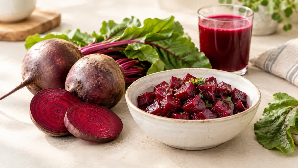 Fresh whole beetroot and sliced beetroot beside a bowl of cooked beetroot, with fresh green leaves and a glass of beetroot juice on a light, neutral surface.