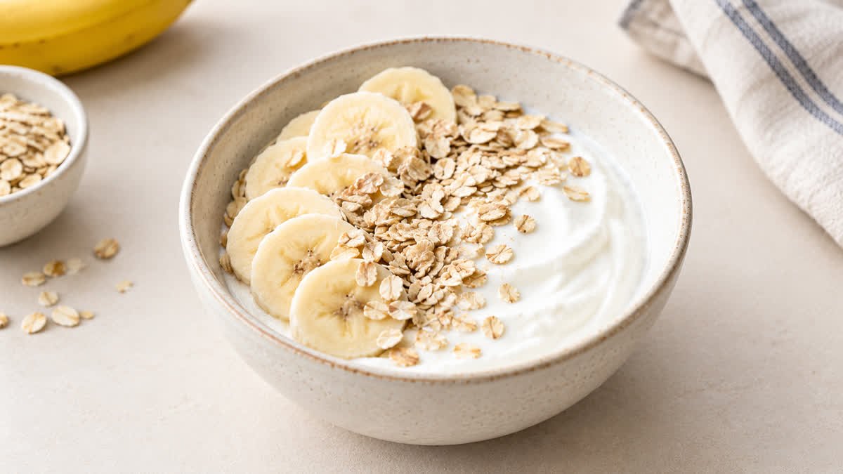 Bowl of plain Greek yogurt with banana slices and oats on a light-colored table.