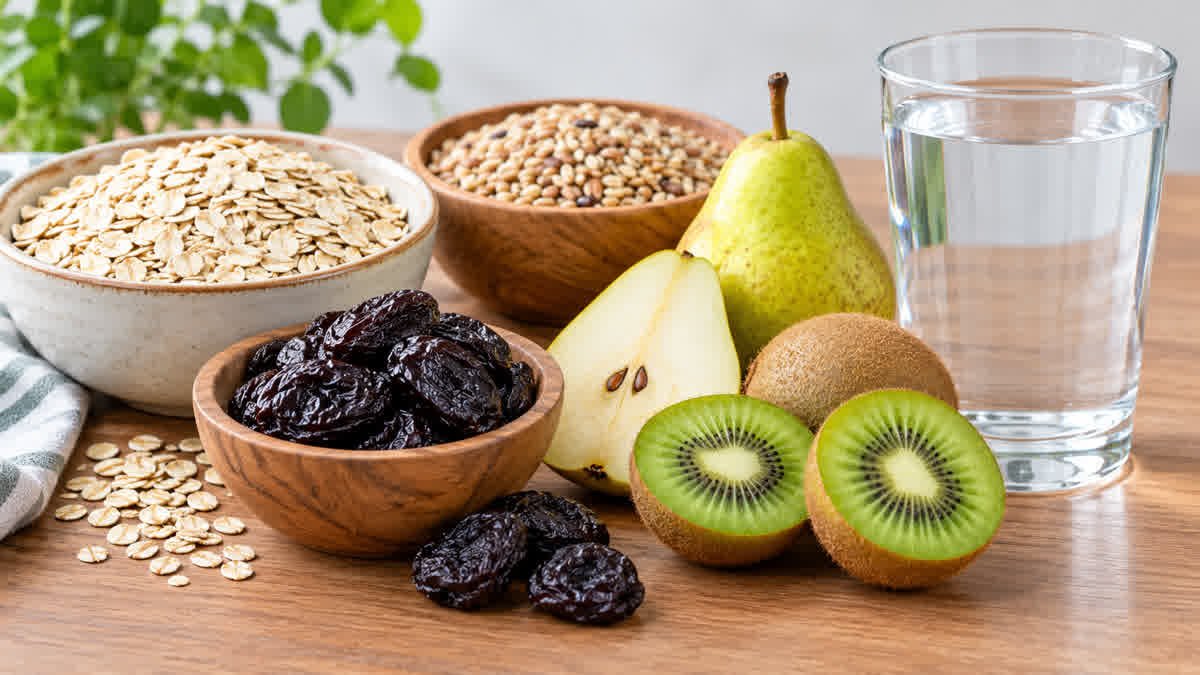 Fiber-rich foods including oats, pears, kiwifruit, and prunes with a glass of water for constipation self-care.
